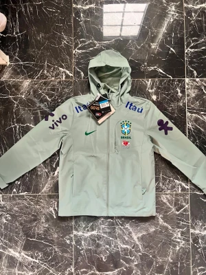Grön Brasilien vindjacka Nike - Snygg grön vindjacka från Nike med Brasilien CBF-logga och flera sponsortryck som Itaú, Vivo och Guaraná Antarctica. Jackan har huva, hel dragkedja och är tillverkad i GORE-TEX-material. Perfekt för dig som gillar fotboll och streetwear.