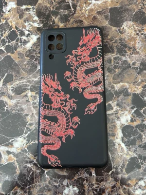 Svart mobilskal med japanska drakar Samsung A12 - Snyggt svart mobilskal med två detaljerade röda drakar på baksidan. Skalet är gjort i mjukt plastmaterial och har utskärningar för kamera och knappar. Perfekt för dig som vill ge mobilen en cool och unik look. För Samsung A12