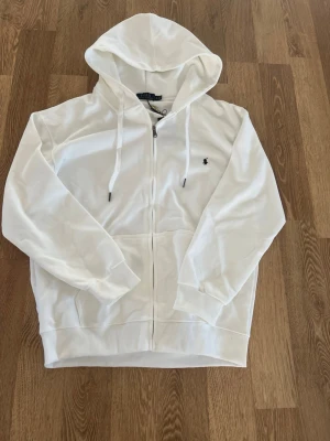 Vit hoodie från Polo Ralph Lauren - Snygg vit hoodie med dragkedja från Polo Ralph Lauren. Klassisk design med huva och dragsko, samt broderad logga på bröstet. Mjuk bomullskänsla och känguruficka framtill. Perfekt för en clean och stilren look.