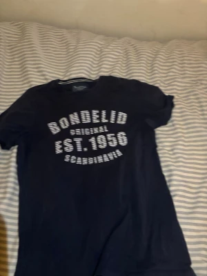 Svart Bondelid t-shirt med tryck - Snygg svart t-shirt från Bondelid med vitt tryck på bröstet där det står 'Bondelid Original EST. 1956 Scandinavia'. Klassisk passform och rund hals. Perfekt till jeans eller shorts för en avslappnad look. (Passar S också)