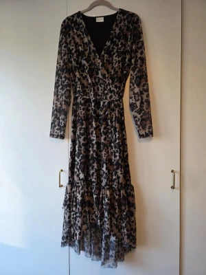 Leopardmönstrad långklänning från Vila - Snygg långklänning från Vila med leopardmönster i svart och beige toner. Klänningen har v-ringning / omlottringning, långa ärmar och volangdetalj nertill. Med skärp en häla trasig men man kan knyta runt midjan i alla fall. Tillverkad i ett lätt och luftigt polyestertyg som ger ett fint fall. 