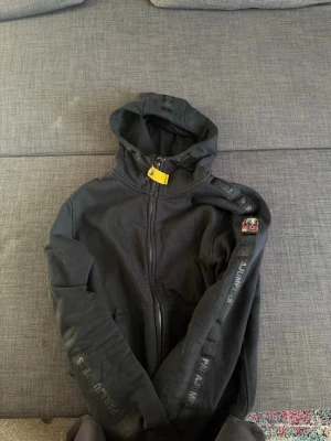 Svart hoodie från Parajumpers - Svart hoodie från Parajumpers med dragkedja, huva och fleecefoder. Märkets logga på ärmen och tryck längs ärmarna. Praktiska fickor framtill och snygga detaljer som gul rem vid halsen. Perfekt för en chill och stilren look. Priset kan diskuteras .