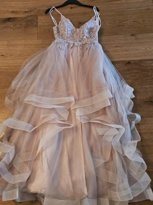 Ljusrosa långklänning med spetsdetaljer - Romantisk långklänning i ljusrosa med voluminös tyllkjol och vackert broderat liv. Klänningen har smala axelband, v-ringning och flera lager volanger för extra dramatik. Perfekt för bal eller speciella tillfällen.