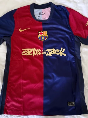 Barça x Cactus Jack matchtröja Nike - Barcelona matchtröja i rött och marinblått med gul Nike-logga och klubbmärke. Framsidan har Cactus Jack-tryck och baksidan UNHCR-logga. Korta ärmar, sportig passform och tillverkad i lätt polyester. Perfekt för fotboll eller streetwear.