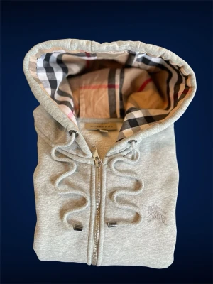 Grå zip hoodie från Burberry - Snygg grå hoodie från Burberry med dragkedja och klassiskt rutigt foder i huvan. Hoodien har dragsko i huvan, ribbade muddar och diskret Burberry-logga broderad på bröstet. Perfekt för dig som gillar stilrena och exklusiva plagg. Står stolen S men är mer som en storlek M i passformen! Dragkedjan är sönder och den är lite använd därför så billigt pris 