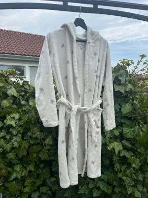 Vit morgonrock med stjärnor och huva - Mysig vit morgonrock med grå stjärnor, huva och knytskärp i midjan. Modellen har två stora fickor framtill och är tillverkad i ett mjukt, fluffigt material som känns skönt mot huden. Perfekt för lata morgnar eller kvällar hemma.