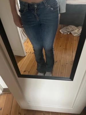 Blå bootcut jeans med fickdetaljer - Säljer ett par blå bootcut jeans med snygga fickdetaljer bak och klassisk femficksdesign. Jeansen har normalhög midja och är i ett stretchigt denimtyg som sitter tajt upptill och släpper ut vid bensluten. Perfekta för en avslappnad men trendig look. Har lite slitage på bakfickan, sista bilden, men inget man tänker på.