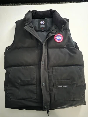 Svart dunväst  - Snygg svart dunväst med hög krage och två stora framfickor. Västen har det klassiska runda Canada Goose-märket på bröstet och dragkedja med tryckknappar. Perfekt för kalla dagar och riktigt street.
