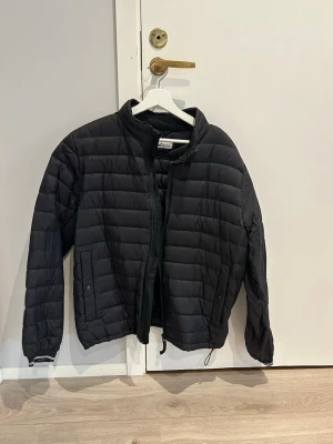 Svart dunjacka från Stone Island - Snygg svart dunjacka från Stone Island med klassisk patch på ärmen. Jackan har quiltad design, hög krage och dragkedja framtill. Perfekt för kalla dagar och riktigt stilren att bära till det mesta.