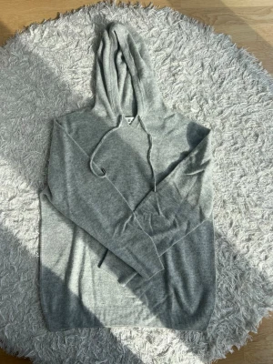 Ljusgrå hoodie i kashmir - En stilren ljusgrå hoodie från GOT i mjukt och tunt stickat kashmir. Klassisk modell med huva och dragsko, samt ribbade muddar vid ärmslut och nederkant. Perfekt för en avslappnad och snygg look. Sitter som en medium.