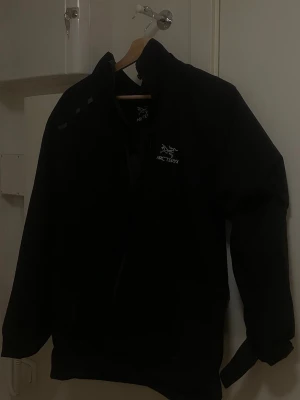 Svart vindjacka från Arc'teryx - Snygg svart vindjacka från Arc'teryx med dragkedja framtill och hög krage. Jackan har minimalistisk design och logga på bröstet. Perfekt för blåsiga dagar och outdoor-aktiviteter. Lätt och smidig att bära med sig. Luva medföljer.