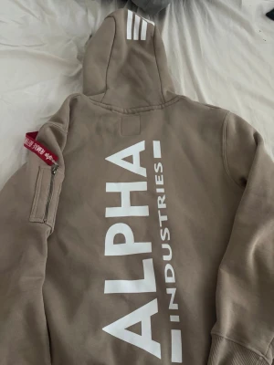 Beige hoodie från Alpha Industries - Snygg beige hoodie från Alpha Industries med stor vit logga på ryggen och ikonisk röd tag på ärmen. Hoodien har huva med tryck och praktisk ficka på ärmen. Perfekt för en streetwear-look.