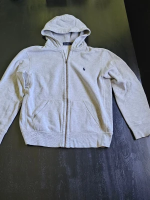 Grå Zip hoodie från Ralph Lauren - Snygg grå hoodie från Ralph Lauren med dragkedja och klassisk broderad logga på bröstet. Tröjan har huva, två fickor framtill och ribbade muddar. Perfekt för en chill och stilren look.