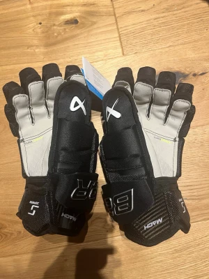 Svarta hockeyhandskar Bauer Supreme mach - Säljer ett par svarta hockeyhandskar från Bauer Supreme mach. Handskarna har grå insida, förstärkta fingrar och handflata samt vaddering för extra skydd. Märkta med 'Mach' och 'Dura' för slitstyrka. Perfekta för dig som spelar ishockey. Aldrig använda. Nypris 2199kr