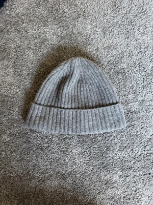 Grå ribbstickad mössa - En stilren grå mössa i ribbstickat material med uppvikt kant. Perfekt för kalla dagar och enkel att matcha med olika outfits. Klassisk form som sitter skönt på huvudet.