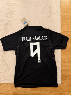Svart Norge fotbollströja Nike - Säljer en svart Norge fotbollströja från Nike med 'BRAUT HAALAND' och nummer 9 på ryggen. Tröjan har korta ärmar, rund hals och är tillverkad i ett lätt, ventilerande material perfekt för träning eller match.