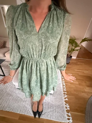 Grön kortklänning med paisleymönster - Snygg grön kortklänning med paisleyliknande mönster och v-ringad hals. Klänningen har långärmade ärmar, resår i midjan och volangig kjol. Perfekt för vår och sommar, tillverkad i ett lätt och luftigt material. Fint skick 