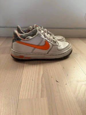 Nike Air Force 1 med orange swoosh - Nike Air Force 1 sneakers i vitt läder med en iögonfallande orange swoosh på sidorna. Skorna har svarta detaljer på hälen och yttersulan, samt klassisk perforerad tå och vita skosnören. Köpta i London.