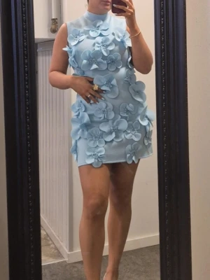 Ljusblå kort klänning med 3D-blommor - Superfin ljusblå kort klänning med stora 3D-blommor framtill och en elegant rosett i nacken. Ärmlös modell med hög hals och figurnära passform. Perfekt för dig som vill sticka ut med en unik och trendig look.