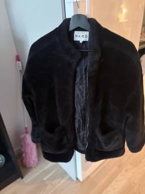 Svart pälsjacka från NA-KD - Mysig svart pälsjacka från NA-KD med kortare modell och rundad krage. Jackan har två fickor framtill och stängs med dragkedja. Perfekt för att hålla värmen under kalla dagar. Tillverkad i mjuk fuskpäls för en trendig look.