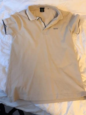 Beige pikétröja från BOSS - Klassisk beige pikétröja från BOSS med korta ärmar och krage. Tröjan har knappar vid halsen och diskret BOSS-logga på bröstet. Perfekt för en stilren och avslappnad look.