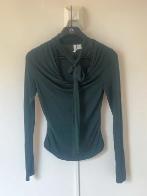 Mörkgrön långärmad topp med knyt - Säljer en mörkgrön långärmad topp från Divided med snygg knytning framtill vid halsen. Toppen har en draperad krage och är figurnära i passformen. Perfekt för dig som gillar stilrena detaljer och mjuka material. Mycket skön kvalitet 