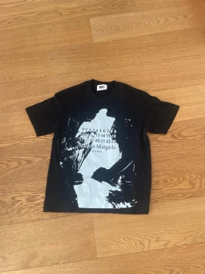Svart t-shirt Maison Margiela - Cool svart t-shirt från Maison Margiela med vit grafisk print och logga på framsidan. Klassisk passform med rund hals och korta ärmar. Ikoniska vita stygn på ryggen. Perfekt för dig som gillar streetwear och designerplagg.