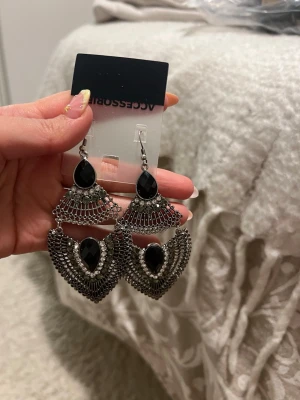 Svarta statement-örhängen Accessorize - Stora, hängande örhängen från Accessorize i silverfärgad metall med svarta stenar och detaljerad design. Örhängena har en unik form med flera lager och gnistrande svarta inslag som ger en cool och trendig look.