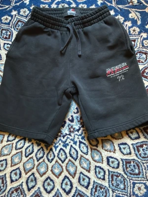 Svarta shorts från McKenzie - Svarta shorts från McKenzie med broderad logga och text på vänster ben. Mjuk resår i midjan med snörning och två sidofickor. Perfekta för en avslappnad stil.