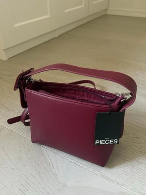 Vinröd handväska från Pieces - Snygg och absolut nytt vinröd handväska från Pieces i slät konstläder med bred axelrem och dragkedja upptill. Väskan har en stilren, fyrkantig form och en liten dekorativ detalj på sidan. Perfekt för dig som vill ha en trendig accessoar med plats för det viktigaste. Pris på prislappen 500:-