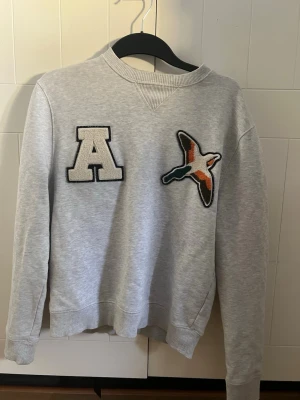 Grå sweatshirt Axel Arigato - Säljer en grå sweatshirt från Axel Arigato med ett stort A-märke och en broderad fågel på bröstet. Tröjan har ribbade muddar vid ärmslut och nederkant samt en avslappnad passform. Perfekt för en casual och trendig look.