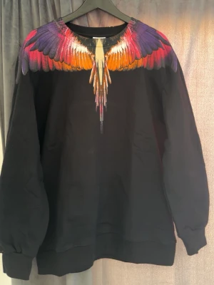 Svart sweatshirt med färgglada vingar - Cool svart sweatshirt från Marcelo Burlon med ett unikt tryck av färgglada vingar i lila, rött, orange och gult över axlar och halsringning. Tröjan har rund hals, långa ärmar och ribbade muddar. Perfekt statement-plagg för dig som vill sticka ut.
