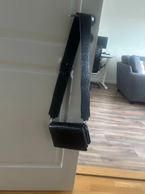 Svart axelväska med bred rem - Snygg svart axelväska i konstläder med bred, justerbar axelrem. Väskan har en stilren, fyrkantig form och stängs med tryckknapp. Perfekt för dig som vill ha en enkel men trendig accessoar till din outfit.