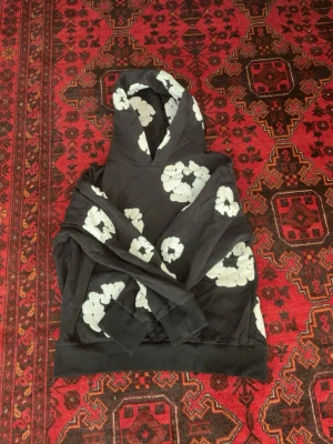 Denim Tears hoodie - Säljer en svart hoodie med stora vita blommönster över hela plagget. Hoodien har en klassisk huva och ribbade muddar vid ärmslut och nederkant. Perfekt för dig som vill sticka ut med en unik och trendig stil.