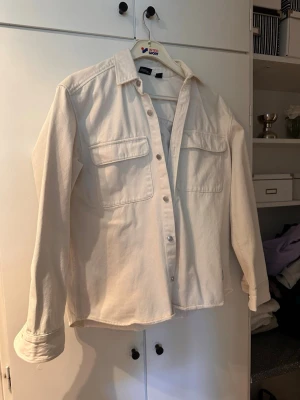 Offwhite overshirt med fickor - Snygg overshirt från Weekday i offwhite bomullstyg. Modellen har raka linjer, två stora bröstfickor med lock och knäppning framtill. Perfekt att slänga över en t-shirt för en avslappnad look.
