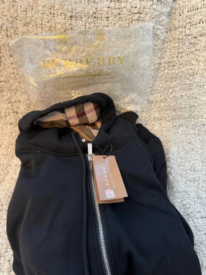 Svart hoodie från Burberry med dragkedja - Helt osnvänd svart hoodie från Burberry med klassiskt rutigt foder i huvan och dragkedja framtill. Märkets logga är diskret broderad på bröstet. Hoodien har en modern passform och är tillverkad i mjukt material för extra komfort.