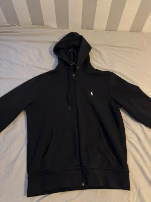 Svart hoodie från Polo Ralph Lauren - Svart hoodie med dragkedja från Polo Ralph Lauren. Klassisk design med vit broderad logga på bröstet, huva med dragsko och två fickor framtill. Perfekt för en chill och stilren look.