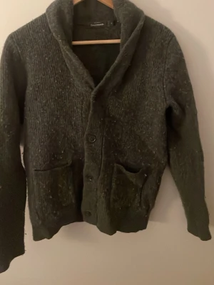 Mörkgrå kofta från Dressmann - Mysig mörkgrå stickad kofta från Dressmann med sjalkrage och två stora fickor framtill. Koftan har knäppning med knappar och är tillverkad i ett grovstickat material som ger en avslappnad look.