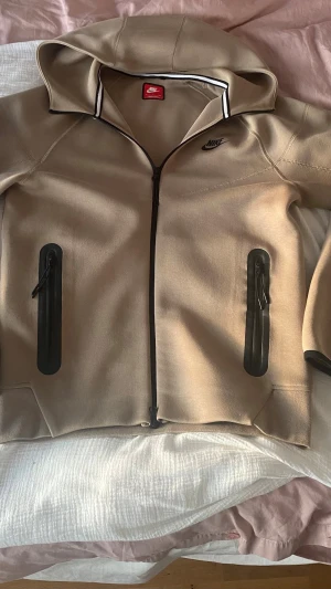 Beige Nike hoodie med dragkedja - Snygg beige hoodie från Nike med hel dragkedja och svarta detaljer. Två fickor med svarta dragkedjor framtill och klassisk Nike-logga på bröstet. Hoodien har en huva och sportig design, perfekt för en avslappnad streetstyle-look.