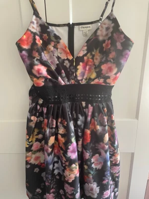 Blommig klänning från Bonprix - Säljer en blommig klänning från Bonprix med v-ringad omlottdesign och smala axelband. Klänningen har ett svart midjeband med spetsdetalj och färgstarkt blommönster i rosa, rött, gult och vitt på mörk botten. Perfekt för sommarens alla tillfällen!
