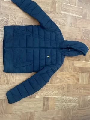 Svart pufferjacka från Lyle & Scott - Säljer en svart pufferjacka från Lyle & Scott med huva och dragkedja. Jackan har två stora fickor framtill och ett diskret gult logomärke på bröstet. 