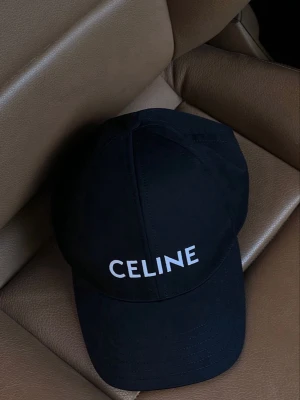 Svart keps från Celine - Stilren svart keps från Celine med vit logotyp broderad framtill. Klassisk böjd skärm och justerbar baksida. Tillverkad i ett mjukt och slitstarkt tyg som ger en clean och modern look.