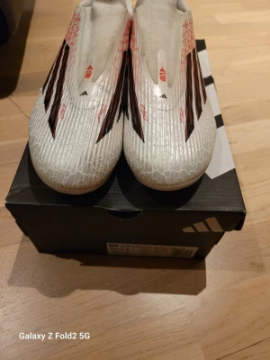 Lamine Yamal vita fotbollsskor F50 LEAGUE LL GG/MG - Säljer ett par vita Lamine Yamal adidas fotbollsskor med svarta och röda detaljer. Skorna har en strömlinjeformad design utan snörning och mönstrad ovandel med blommönster. Perfekta för snabba spelare som vill sticka ut på planen.