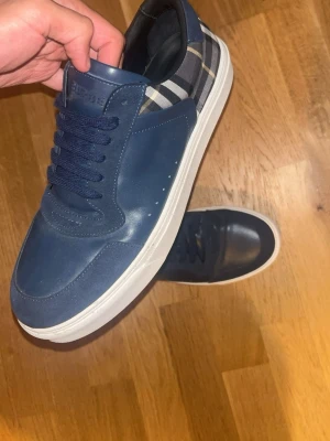 Marinblå sneakers från Burberry - Säljer ett par marinblå sneakers från Burberry med klassiskt rutigt mönster på hälen. Skorna har vita sulor, mörkblå snören och är tillverkade i läder med textildetaljer. Perfekta för dig som gillar stilrena och exklusiva skor med en twist.