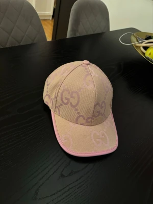 Beige och rosa keps från Gucci - Säljer en snygg keps från Gucci i rosa färg och GG-logga över hela kepsen (Jumbo model). Kepsen har böjd skärm och rosa kant runt om. Perfekt till sommaren!
