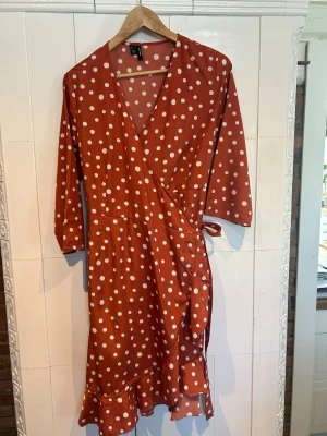 Rostbrun omlottklänning med prickar Vero Moda - Snygg röd omlottklänning från Vero Moda med vita prickar och volang nertill. Klänningen har trekvartsärm och knyt i midjan för en smickrande passform.
