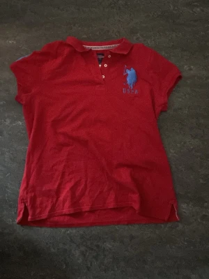Röd pikétröja från U.S. Polo Assn. - Röd pikétröja från U.S. Polo Assn. med klassisk krage, korta ärmar och blå logga broderad på bröstet. Tröjan har knappar vid halsen och är tillverkad i mjuk bomull. Perfekt för en sportig och avslappnad stil. Storlek xl känns dock som M-L pris kan också diskuteras.