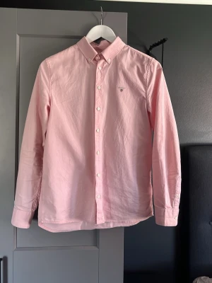 Ljusrosa skjorta från GANT - Snygg ljusrosa skjorta från GANT med klassisk button-down krage och vita knappar. Skjortan har lång ärm och liten broderad logga på bröstet. Perfekt för dig som gillar stilrena plagg med en fräsch färg. Skjortan passar någon som är runt 170 cm, är 174 själv och den är lite kort överlag, vid frågor är det bara att skriva!