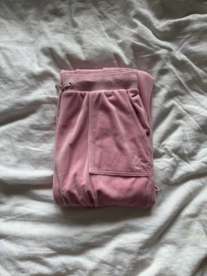Rosa mjukisbyxor från Juicy Couture - Supermjuka rosa mjukisbyxor från Juicy Couture i velour med broderad JC-logga på benet och bred resår i midjan med dragsko. Perfekta för en chill och avslappnad stil.