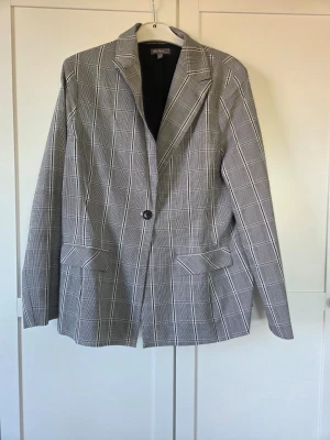 Grårutig kavaj, stl 46, Ulla popken  - Snygg grå kavaj med vitt rutmönster och klassisk krage. Perfekt för en trendig och avslappnad look. Materialet känns mjukt och följsamt.
